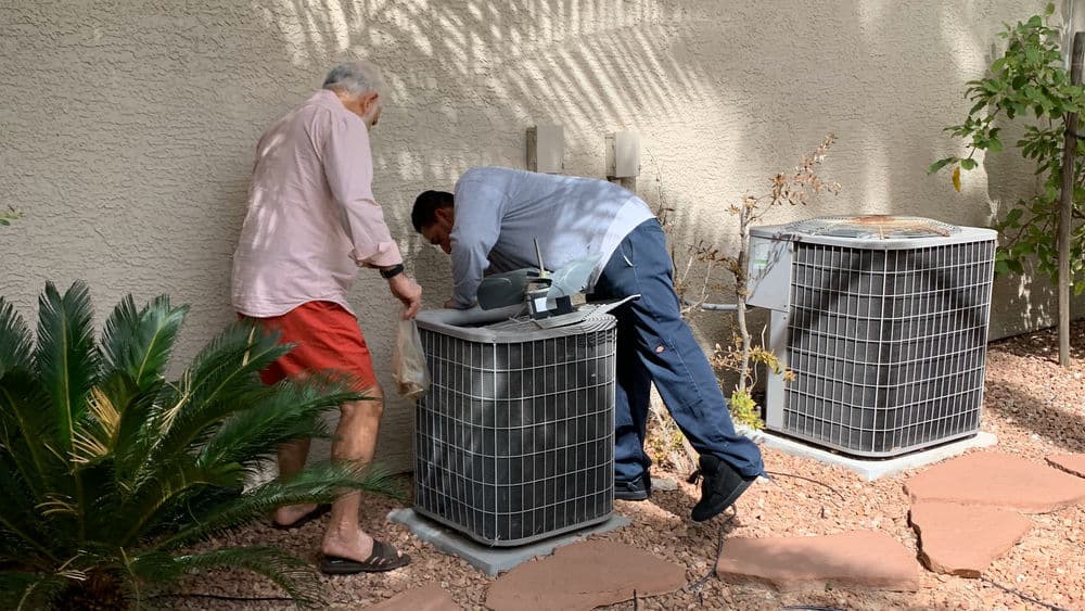 Two technicians inspecting air conditioning units outdoors near a house.