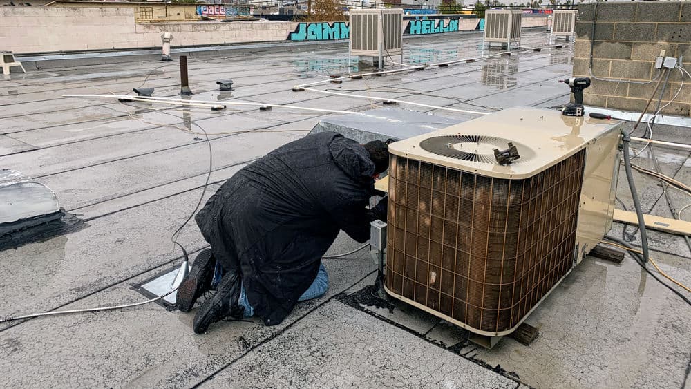 Technician performing maintenance on rooftop air conditioning unit during rainy weather.